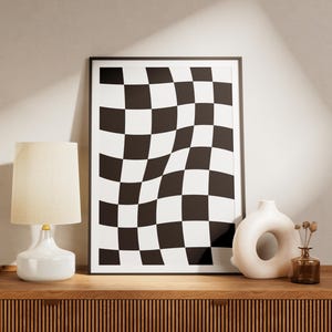 May include: A framed black and white checkered art print with a wavy design. A white ceramic lamp with a beige lampshade and a white vase with a hole are on a wooden surface.
