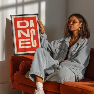 May include: A framed art print with the word "DANCE" in white block letters on a red background. The print is held by a person wearing a denim jacket and pants, sitting on an orange sofa. The room has natural light.