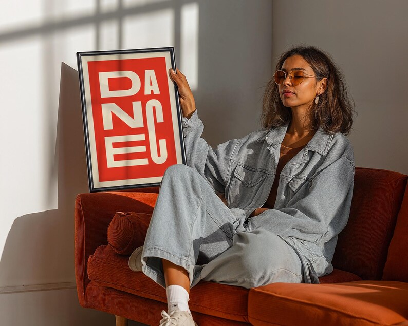 May include: A framed art print with the word "DANCE" in white block letters on a red background. The print is held by a person wearing a denim jacket and pants, sitting on an orange sofa. The room has natural light.