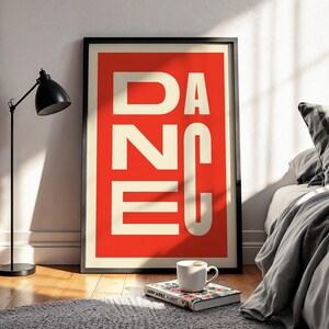 May include: A framed art print with the word "DANCE" in large white capital letters against a red background. The print is displayed on a wooden floor, leaning against a white wall. A black lamp, a book, and a coffee cup are in the foreground.