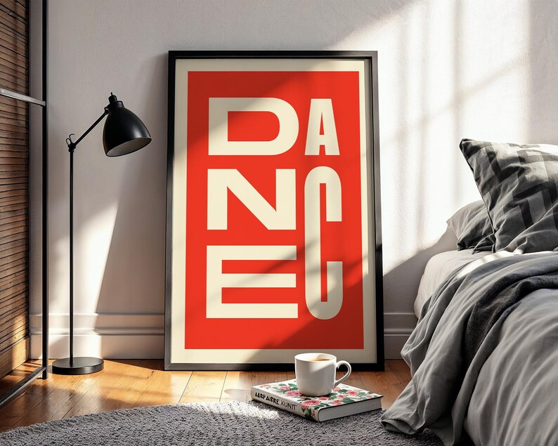 May include: A framed art print with the word "DANCE" in large white capital letters against a red background. The print is displayed on a wooden floor, leaning against a white wall. A black lamp, a book, and a coffee cup are in the foreground.