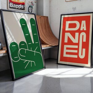 May include: Two framed art prints. One features a green peace sign hand gesture on a cream background. The other print has the word "DANCE" in white block letters on a red background. Both are in black frames.