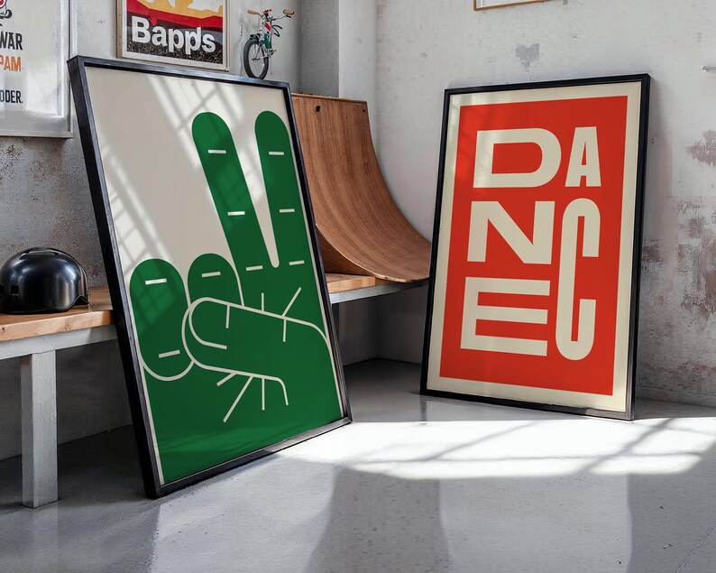 May include: Two framed art prints. One features a green peace sign hand gesture on a cream background. The other print has the word "DANCE" in white block letters on a red background. Both are in black frames.