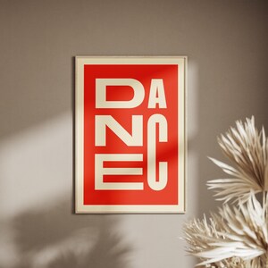 May include: A framed art print with the word "DANCE" in bold, white capital letters against a red background. The frame is a light beige color. The print is hanging on a neutral-toned wall, with dried plants in the corner.