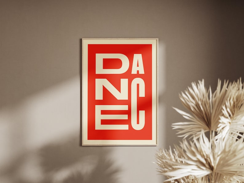 May include: A framed art print with the word "DANCE" in bold, white capital letters against a red background. The frame is a light beige color. The print is hanging on a neutral-toned wall, with dried plants in the corner.
