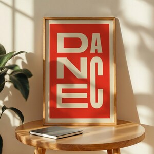 May include: A framed art print with the word "DANCE" in large, cream-colored block letters against a red background. The print is in a light wood frame and rests on a round wooden table. A closed notebook sits below the print.