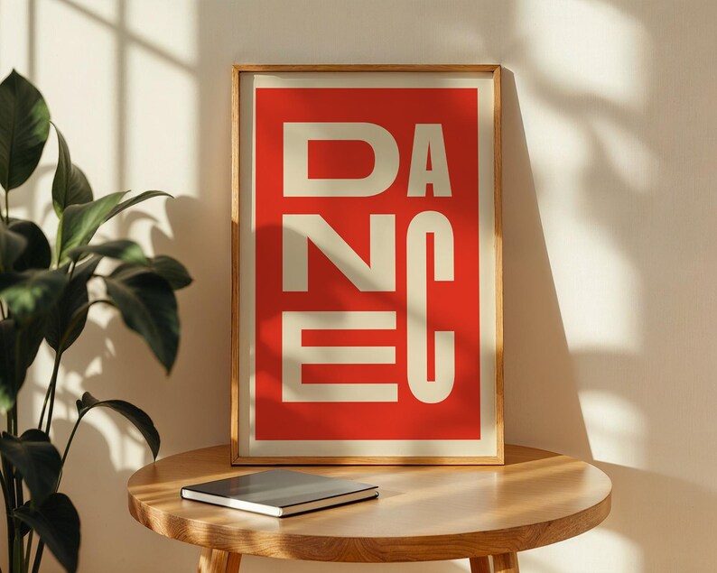 May include: A framed art print with the word "DANCE" in large, cream-colored block letters against a red background. The print is in a light wood frame and rests on a round wooden table. A closed notebook sits below the print.