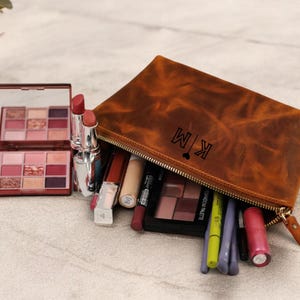 May include: A brown leather zippered pouch with the initials "KM" and a heart symbol embossed on the front. The pouch is open and filled with makeup items, including lipstick, eyeshadow palettes, and eyeliner.