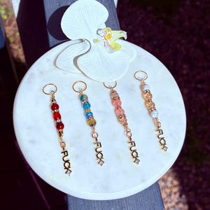 Puede incluir: Cuatro llaveros con cuentas con herrajes dorados y la palabra "FUCK" escrita en letras doradas. Las cuentas son rojas, azules, rosas y naranjas. Los llaveros se exhiben sobre una superficie de mármol blanco con una flor de orquídea blanca.