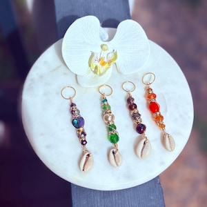 Può includere: Quattro accessori per capelli con perline, cerchi dorati e ciondoli a conchiglia. Le perline sono di vari colori, tra cui viola, verde, marrone e arancione. Un fiore di orchidea bianca su una superficie di marmo bianco.