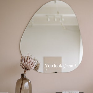 May include: A light brown wooden dresser with a large, irregular shaped mirror hanging on the wall above it. The mirror has the text "You look great" written on it. A vase with a dried pink flower is on the dresser.