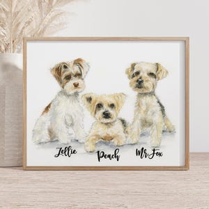 May include: A framed watercolor print featuring three small dogs. The dogs are in shades of white, brown, and tan, with names written below them: Jellie, Peach, and Mr. Fox. The frame is a light wood color.