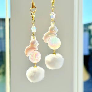 May include: Two decorative charms with gold-colored chains. Each charm features a white bunny, a glittery iridescent ball, and a fluffy white pom-pom. Small star-shaped beads are also included. The charms are hanging against a blurred background.