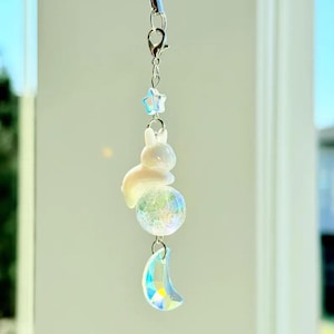 May include: A decorative charm with a white rabbit figure above an iridescent glass ball, and a crystal crescent moon pendant. A small blue star and silver clasp are also present. The charm is suspended by a thin, clear string.