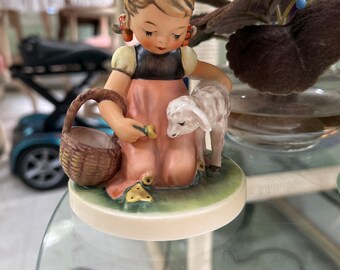 Puede incluir: Figura de cerámica de una niña con vestido rosa, top negro y pelo rubio, sosteniendo una cesta y una flor, con un cordero a su lado. La figura está sobre una base redonda con una combinación de colores verde y marrón.