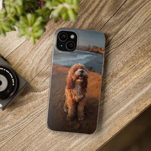 May include: A clear smartphone case featuring a photographic print of a fluffy, golden-brown dog sitting on a sandy beach with the ocean and coastline visible in the background. The case has a cutout for a dual-lens camera system on the back.