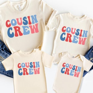 May include: Four cream colored t-shirts with the text "COUSIN CREW" in blue and red letters. The shirts are in different sizes, with one being a baby onesie.