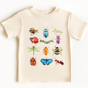 May include: A cream-coloured t-shirt with a colourful insect design. The design features a variety of insects, including butterflies, beetles, and ants, all in watercolour style.