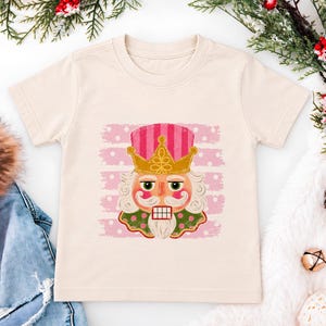Puede incluir: Una camiseta de manga corta color crema con un estampado gráfico de un cascanueces con corona dorada y sombrero de rayas rosas. El cascanueces tiene barba blanca, cuello verde y está centrado sobre un fondo de rayas horizontales rosas.