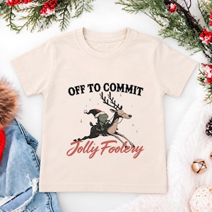 May include: A cream-coloured short-sleeve t-shirt featuring a graphic of a frog wearing a Santa hat riding a reindeer. The text above the graphic reads OFF TO COMMIT and the text below the graphic reads Jolly Foolery in a cursive red font.