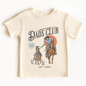 May include: Cream-colored t-shirt featuring a graphic of a cowboy on horseback reaching out to a child cowboy on a horse. The shirt has the text "Dads Club" and "American Made" with the year "2026" and the names "Dad + Henry".