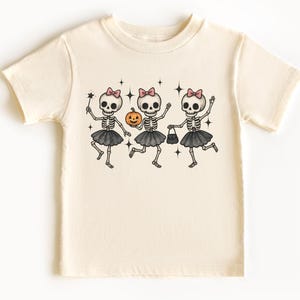 May include: A cream-colored short-sleeve t-shirt featuring a graphic print of three skeletons wearing pink bows and black tutus. The skeletons are depicted with a pumpkin, a small handbag, and a star-shaped wand, surrounded by small star icons.