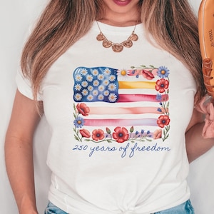 Puede incluir: Camiseta blanca con un dise&ntilde;o de acuarela de una bandera estadounidense adornada con margaritas y amapolas. El dise&ntilde;o de la bandera incluye el texto "250 years of freedom" en una fuente de escritura. Prenda de vestir informal.