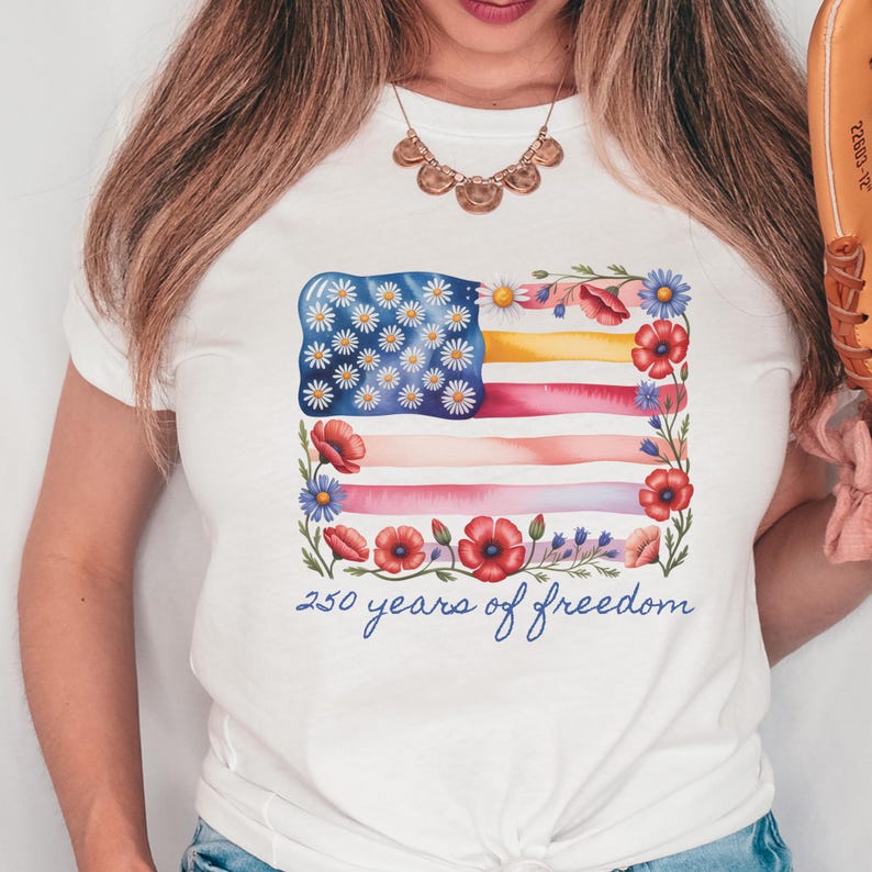 Puede incluir: Camiseta blanca con un dise&ntilde;o de acuarela de una bandera estadounidense adornada con margaritas y amapolas. El dise&ntilde;o de la bandera incluye el texto "250 years of freedom" en una fuente de escritura. Prenda de vestir informal.