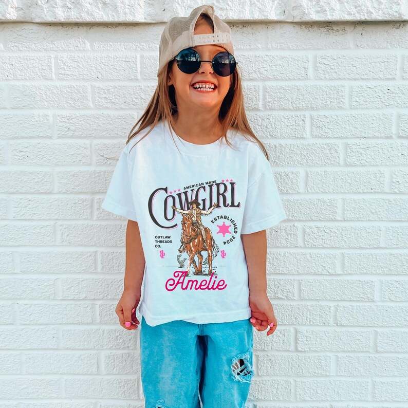 May include: A white t-shirt with a pink and black graphic of a cowgirl riding a horse. The text "Cowgirl" is printed in black with a pink star. The text "Established 2001" is printed in pink. The text "Outlaw Threads Co." is printed in black. The text "Ameli&eacute;" is printed in pink.
