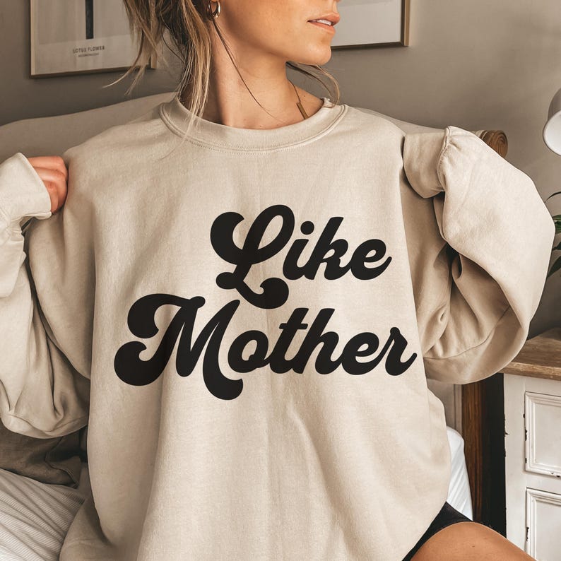 May include: A beige sweatshirt with the words "Like Mother" in large, black, retro-style lettering. The sweatshirt has a crew neck and long sleeves. The image is a close-up shot of the sweatshirt.