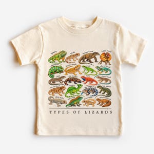 May include: A cream-colored t-shirt featuring a colorful graphic print of various lizard species arranged in a grid. Each lizard is labeled with its name, and the text "TYPES OF LIZARDS" is printed in black capital letters at the bottom.