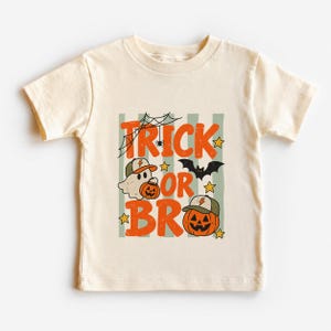 May include: A cream-coloured short-sleeve t-shirt featuring a graphic print with the text "TRICK OR BRO" in orange. The design includes a ghost wearing a baseball cap, a jack-o'-lantern, a black bat, a spider web, and small yellow stars.
