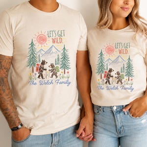 Peut inclure: Un t-shirt à manches courtes de couleur crème avec un motif représentant deux ours en randonnée dans une forêt avec des montagnes et un soleil. Le texte indique "LET'S GET WILD." et "The Welch Family" en bleu. Le design inclut des arbres verts.