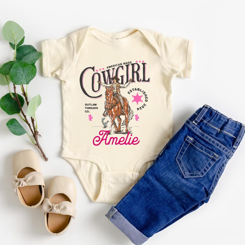 May include: A cream colored baby onesie with a pink and black graphic of a cowgirl riding a horse. The graphic includes the text "Cowgirl", "American Made", "Outlaw Threads Co.", and "Established 2024". The onesie is personalized with the name "Amelie".