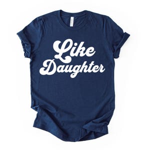 May include: Navy blue t-shirt with the words "Like Daughter" in white, cursive lettering. The shirt has rolled-up sleeves and a classic crew neck. The shirt is made of a soft material.