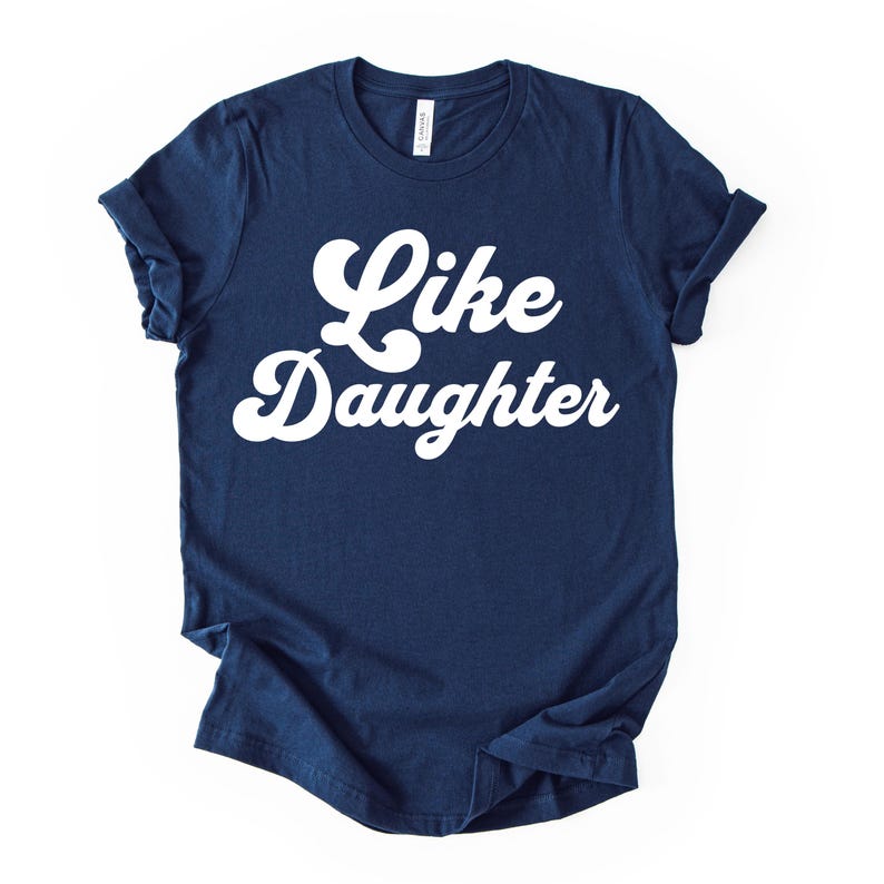 May include: Navy blue t-shirt with the words "Like Daughter" in white, cursive lettering. The shirt has rolled-up sleeves and a classic crew neck. The shirt is made of a soft material.