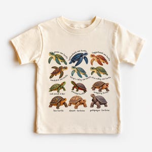 Puede incluir: Una camiseta de manga corta de color crema con una cuadrícula de doce ilustraciones detalladas de varias tortugas, cada una etiquetada con su nombre común, incluyendo la tortuga verde, la tortuga laúd, la tortuga boba y la tortuga mordedora.