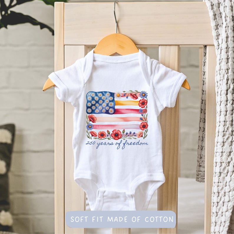 Puede incluir: Body de beb&eacute; blanco con un dise&ntilde;o de acuarela de la bandera estadounidense. La bandera presenta margaritas azules y blancas, rayas rojas y rosas y un borde floral. El texto "250 years of freedom" est&aacute; debajo.