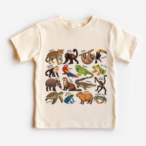 May include: A cream-colored short-sleeve t-shirt featuring a colorful graphic print of various rainforest animals including a jaguar, sloth, toucan, macaw, iguana, caiman, capybara, and sea turtle, each labeled with its name in black text.