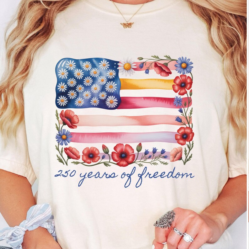 Puede incluir: Camiseta color crema con un dise&ntilde;o floral de la bandera estadounidense. Las estrellas de la bandera est&aacute;n representadas por margaritas blancas, y las rayas son en tonos acuarela. Amapolas rojas y flores azules enmarcan la bandera, con el texto "250 years of freedom" en azul.