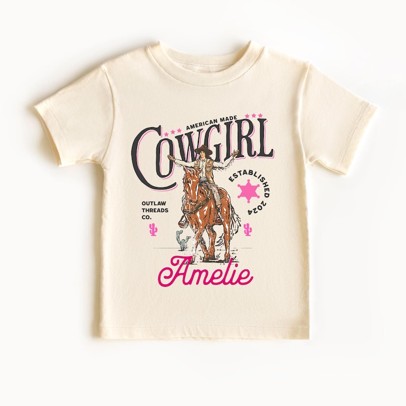 May include: A cream colored t-shirt with a vintage graphic of a cowgirl riding a horse. The graphic includes the text "Cowgirl", "American Made", "Outlaw Threads Co.", and "Established 2024". The name "Amelie" is printed below the graphic in pink.