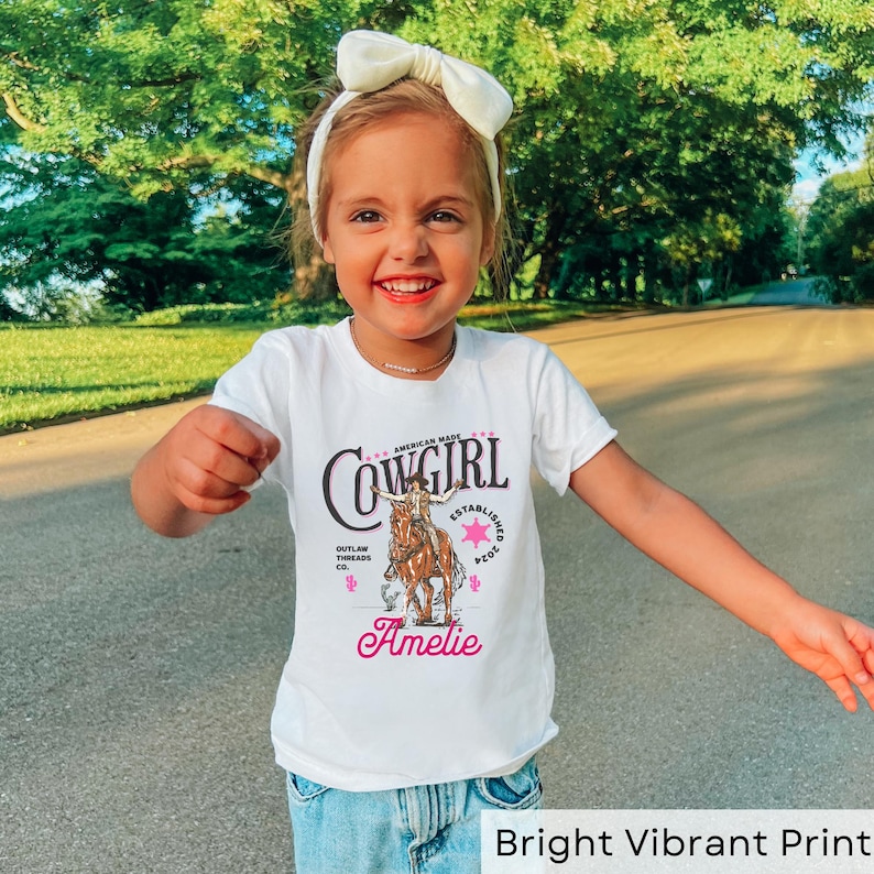 May include: White t-shirt with a pink and black graphic of a cowgirl riding a horse. The graphic includes the text "Cowgirl", "American Made", "Established 2002", and "Outlaw Threads Co.". The shirt is personalized with the name "Amelie".