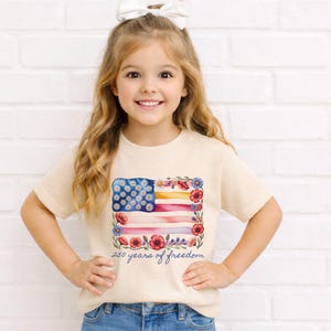 Puede incluir: Una camiseta beige con un dise&ntilde;o floral de la bandera estadounidense. La bandera est&aacute; compuesta por margaritas azules, amapolas rojas y rayas amarillas y rosas. El texto "250 years of freedom" est&aacute; impreso debajo.