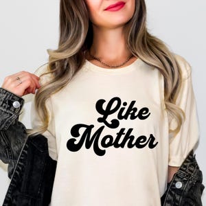 May include: Cream-colored t-shirt with the words "Like Mother" in bold, black, retro-style lettering. The shirt is a crew neck and has short sleeves. The model is wearing a black denim jacket.