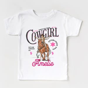May include: White t-shirt with a pink graphic of a cowgirl riding a horse. The graphic includes the text "Cowgirl", "American Made", "Outlaw Threads Co.", and "Established 2024". The name "Amelie" is written in pink cursive below the graphic.