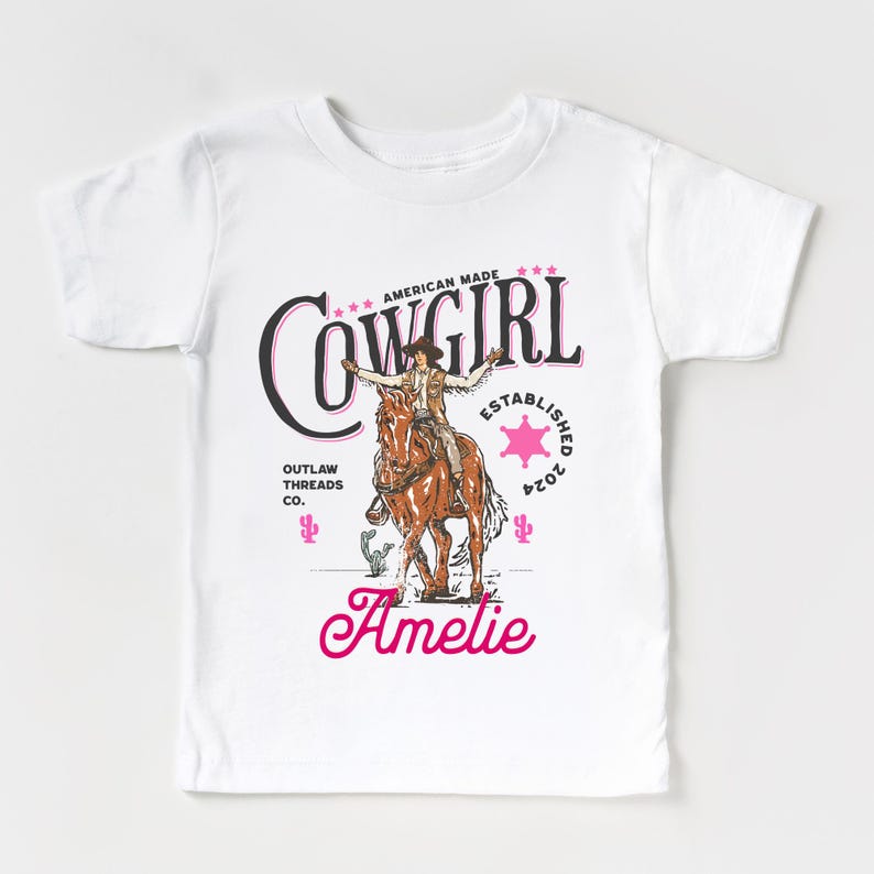May include: White t-shirt with a pink graphic of a cowgirl riding a horse. The graphic includes the text "Cowgirl", "American Made", "Outlaw Threads Co.", and "Established 2024". The name "Amelie" is written in pink cursive below the graphic.