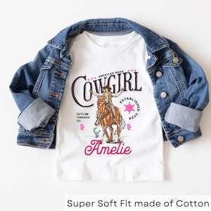 May include: White t-shirt with a pink and black graphic of a cowgirl riding a horse. The graphic includes the text "American Made Cowgirl Established 2024 Outlaw Threads Co.". The name "Amelie" is written in pink below the graphic.