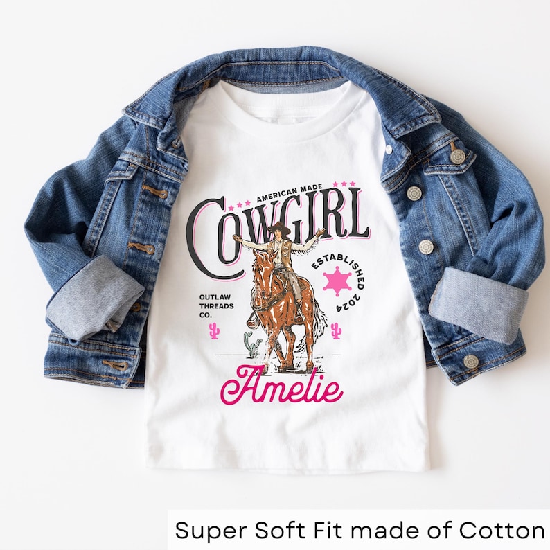 May include: White t-shirt with a pink and black graphic of a cowgirl riding a horse. The graphic includes the text "American Made Cowgirl Established 2024 Outlaw Threads Co.". The name "Amelie" is written in pink below the graphic.
