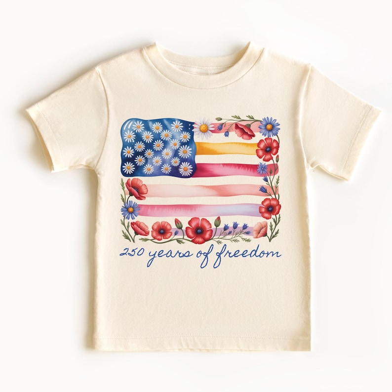 Puede incluir: Camiseta de color crema con un dise&ntilde;o de acuarela de la bandera estadounidense adornada con flores. El dise&ntilde;o de la bandera incluye margaritas, amapolas y otros elementos florales. El texto "250 years of freedom" est&aacute; escrito en gui&oacute;n azul.