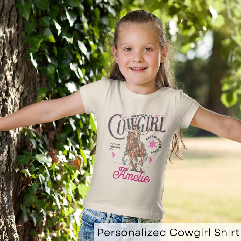May include: A personalized cowgirl t-shirt with a vintage design featuring a cowgirl riding a horse. The shirt is cream colored with pink text that says "American Made Cowgirl Established 2021 Outlaw Threads Co." and "Ameli."